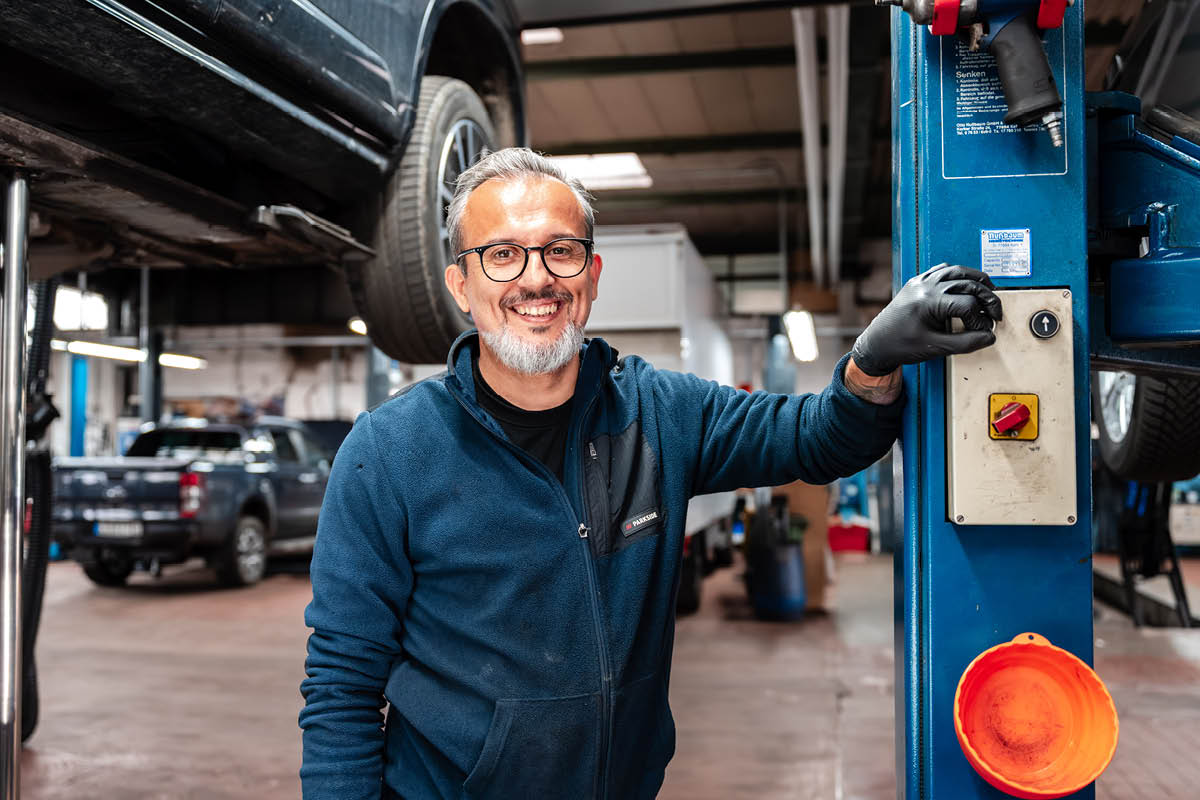 Mechaniker lehnt sich an eine Hebebühne in der Werkstatt und lächelt in die Kamera