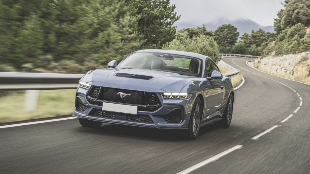 Blauer Ford Mustang GT fährt auf einer kurvenreichen Landstraße, umgeben von Bäumen und Bergen im Hintergrund